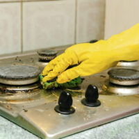 Cleaning dirty gas stove from grease, food leftovers deposits. woman's hand in protective glove with sponge rag and detergent washing kitchen stove. home cleaning service concept.